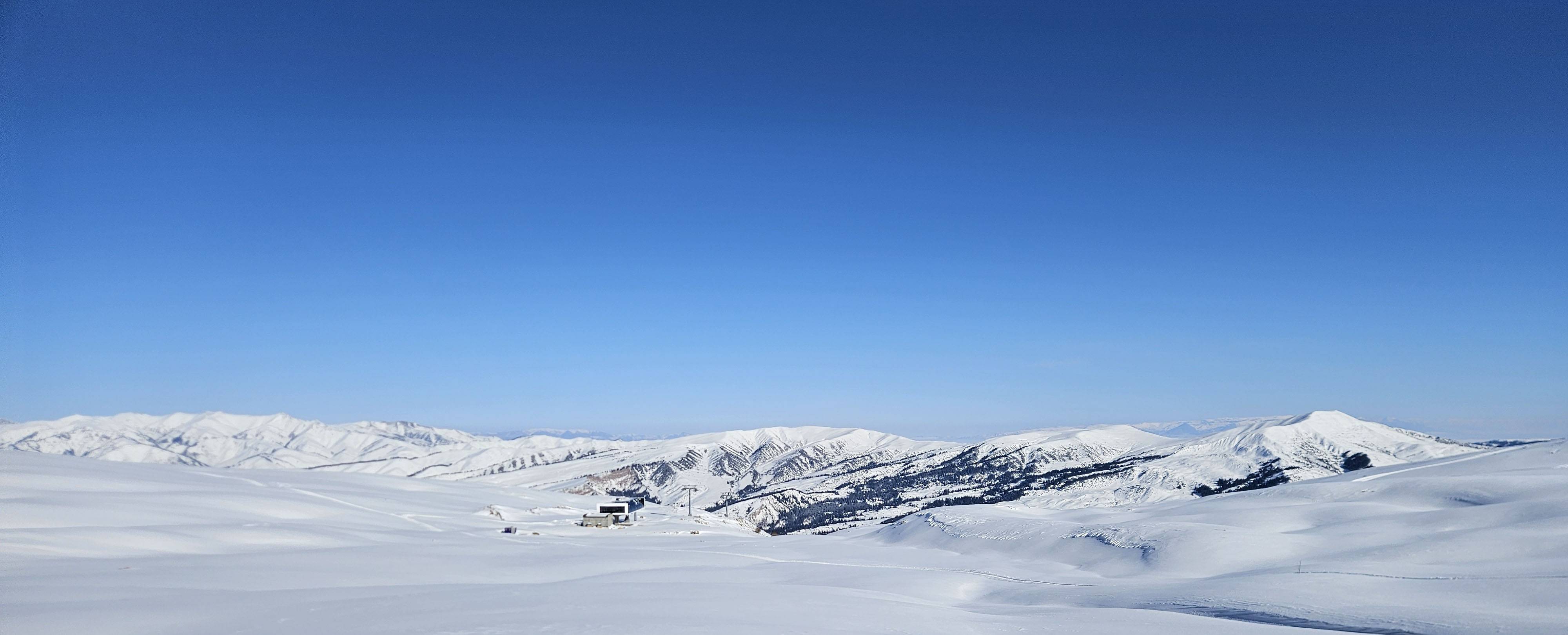 Tief verschneites Hochgelände im Tian Shan unter wolkenlosem Himmel, in der Bildmitte ein Gebäude und Liftstützen