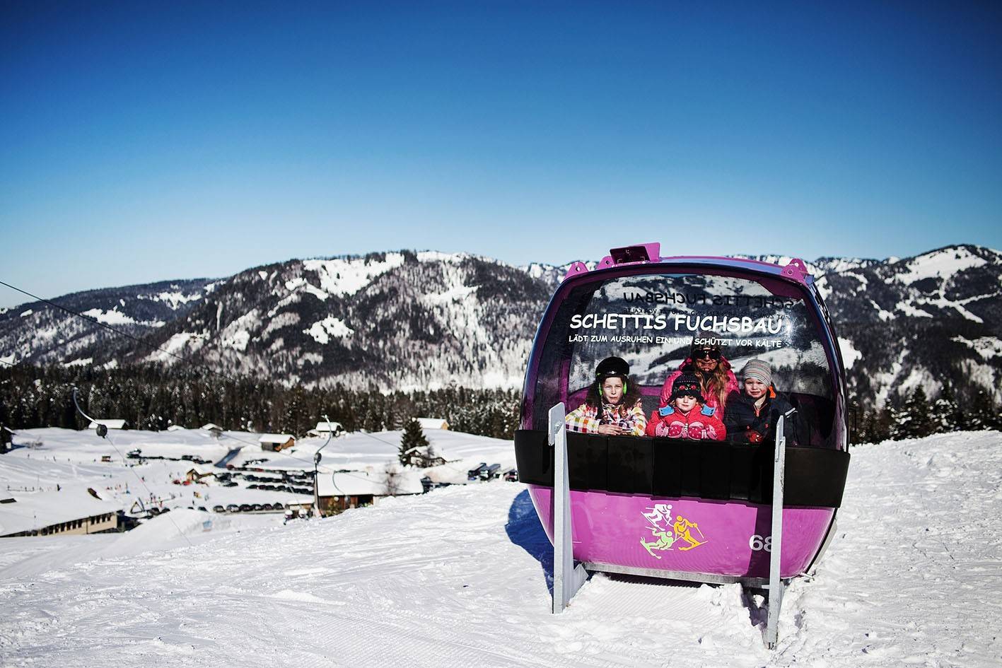 Familie in der pinken Gondelkabine „Schettis Fuchsbau“ im Schnee, dahinter das Bregenzerwälder Bergpanorama